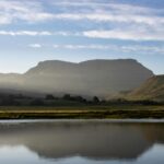 A lake near a mountain under a blue sky with the mountain shadow seen in the waters