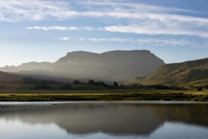 A lake near a mountain under a blue sky with the mountain shadow seen in the waters