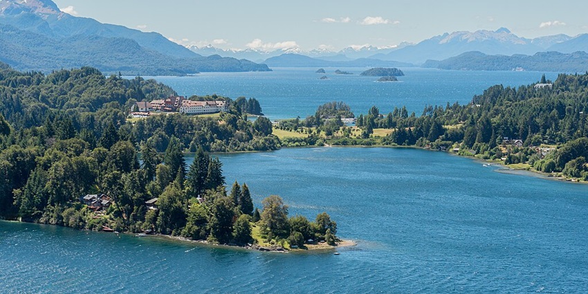 An aerial view of a huge lake with a few houses on the left surrounded by greenery and hills