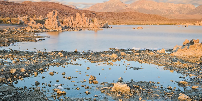 An image of Mono Lake in California with rocky and sandy shores and brown sandy mountains