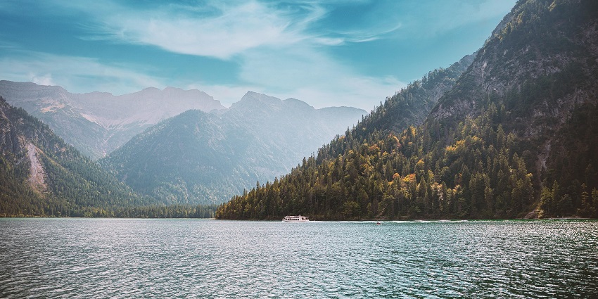 A serene lake surrounded by green mountains under a bright blue sky, with a boat sailing