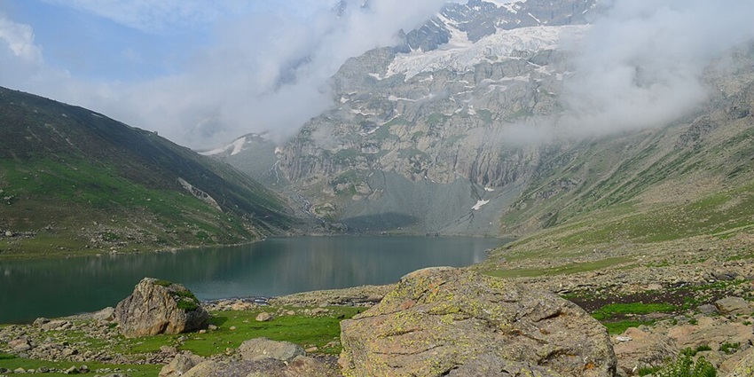 Nundkol Lake with multiple big rocks in the front, green shorelines and mountains