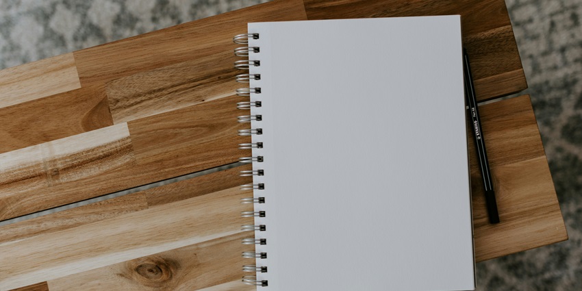 A blank spiral-bound notepad sits open on a rustic wooden table next to a black pen