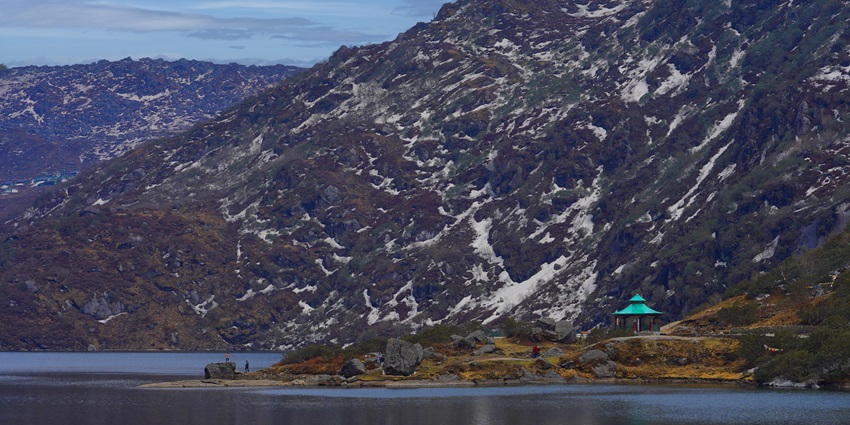 Sandy shore of Tsomgo Lake with big rocks, and people standing on the rocks and mountains