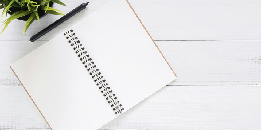 Open spiral notebook with blank pages, a pen, and a potted plant on a white wooden table