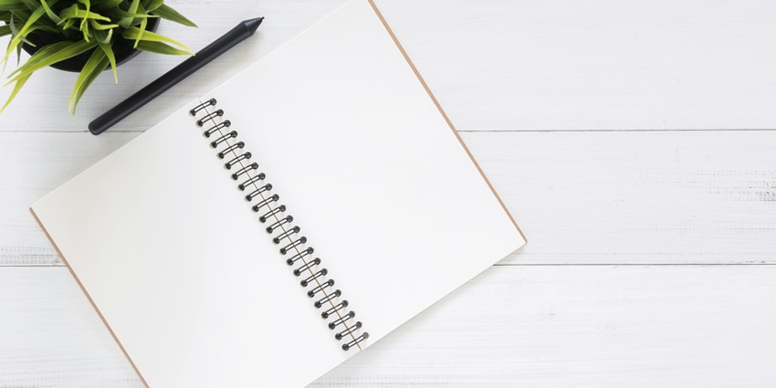 Open spiral notebook with blank pages, a pen, and a potted plant on a white wooden table