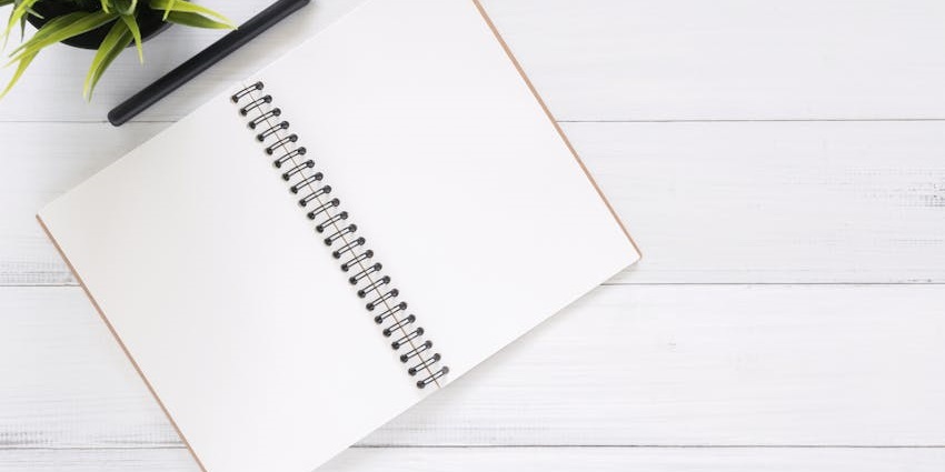 Open spiral notebook with blank pages, a pen, and a potted plant on a white wooden table