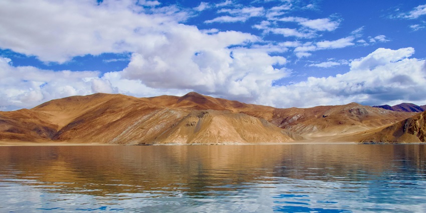 Pangong Tso with a large blue water body, sandy shorelines surrounded by rocky hills.
