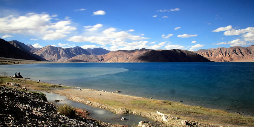 Pangong Tso with stones piled, bright blue water bodies, sandy shorelines and mountains.