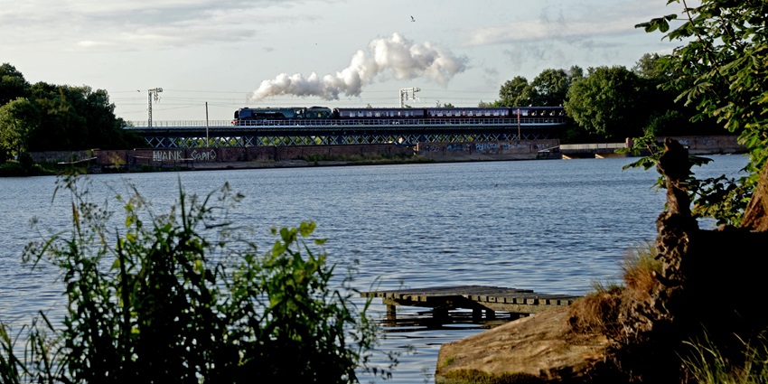 A calm lake with a steam train passing over a bridge in the background