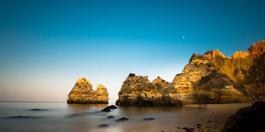 Jagged cliffs and rock formations lining a sandy beach at dusk, with the ocean