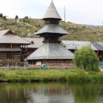 Prashar Lake in summer with Parashar temple, multiple flags and distant stairs leading up the hill.