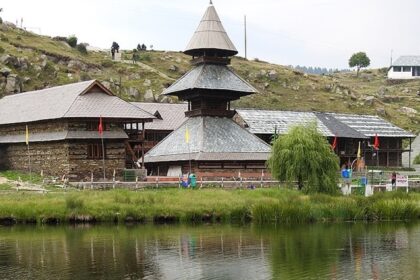 Prashar Lake in summer with Parashar temple, multiple flags and distant stairs leading up the hill.