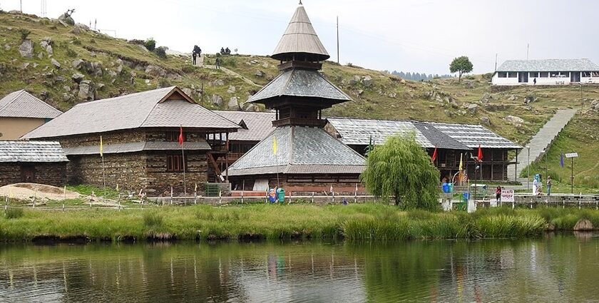 Prashar Lake in summer with Parashar temple, multiple flags and distant stairs leading up the hill.