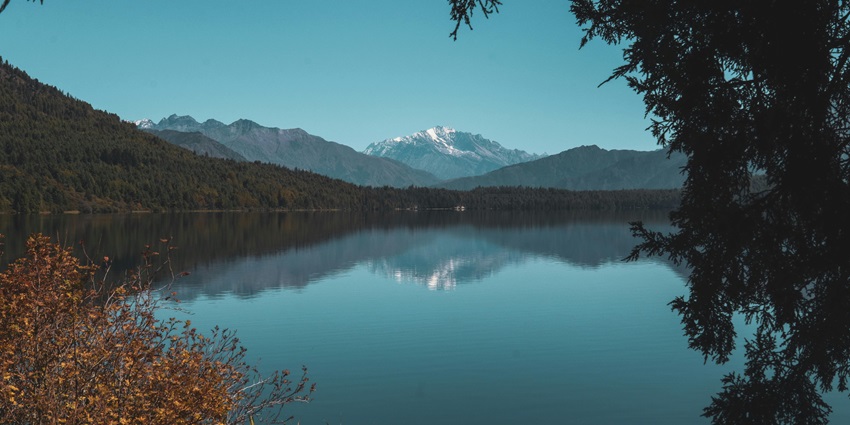 Rara Lake with a wooden bench on the green shoreline, a rainbow forming and green hills.