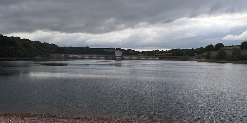 Dam over a lake with a sandy shoreline and small slope-like hills covered in greenery