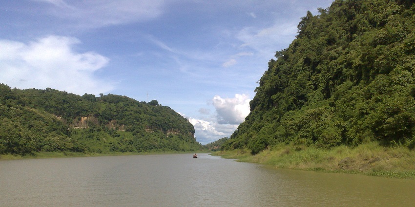Lake with a calm brown water body, big green hills on both sides under a clouded sky