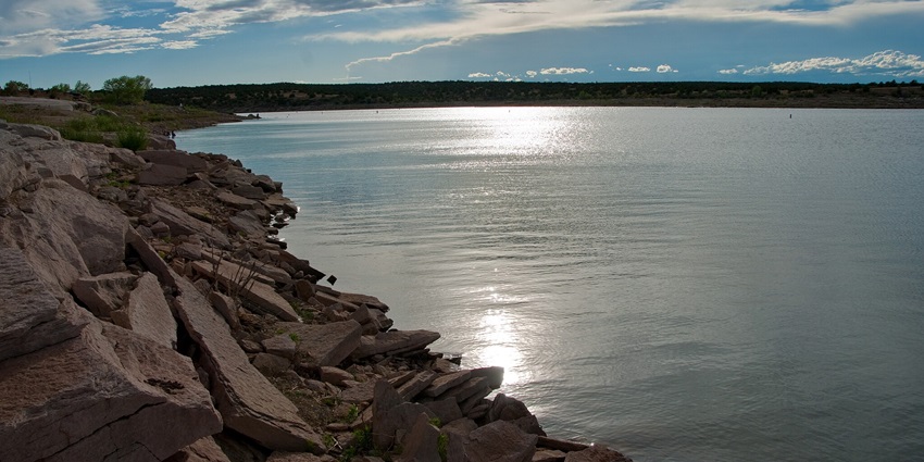 Lake with a rocky shoreline, distant trees and reflection of sun under the clouded sky