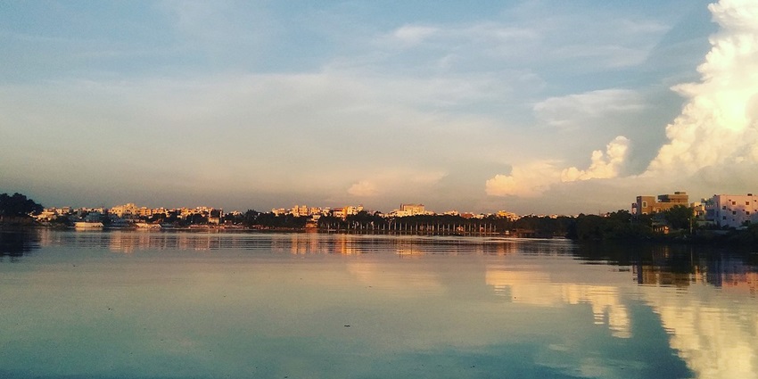 An image of Saroornagar Lake with calm waters, a cityscape, and a pastel sky.
