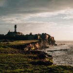 An image of a sea fort of Diu, with a tall lighthouse on grassy cliffs under cloudy sky.