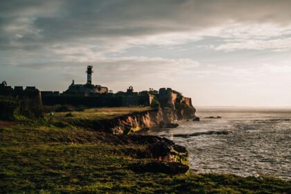 An image of a sea fort of Diu, with a tall lighthouse on grassy cliffs under cloudy sky.