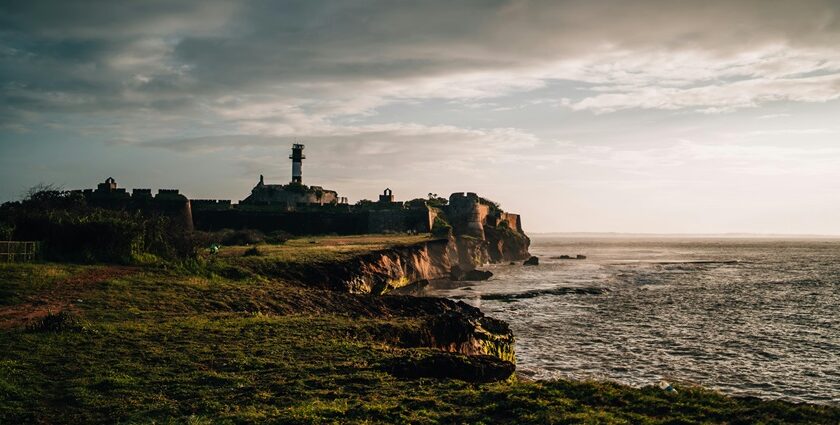 An image of a sea fort of Diu, with a tall lighthouse on grassy cliffs under cloudy sky.