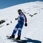 An image of a man on a snow-covered mountain with rugged peaks, preparing for snowboarding in India.