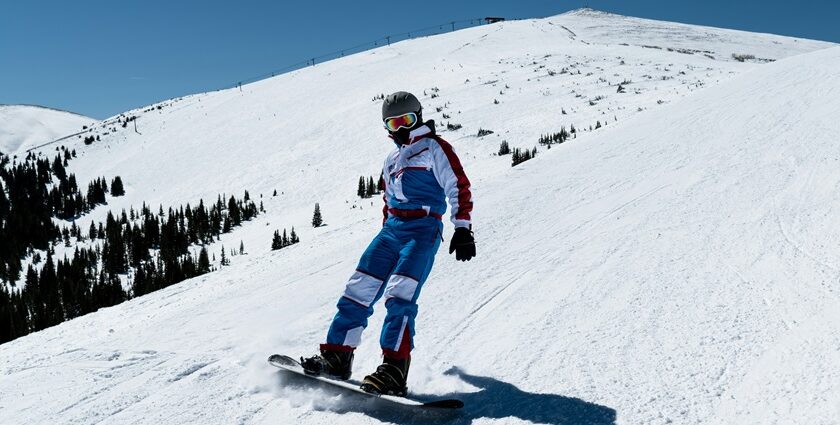 An image of a man on a snow-covered mountain with rugged peaks, preparing for snowboarding in India.