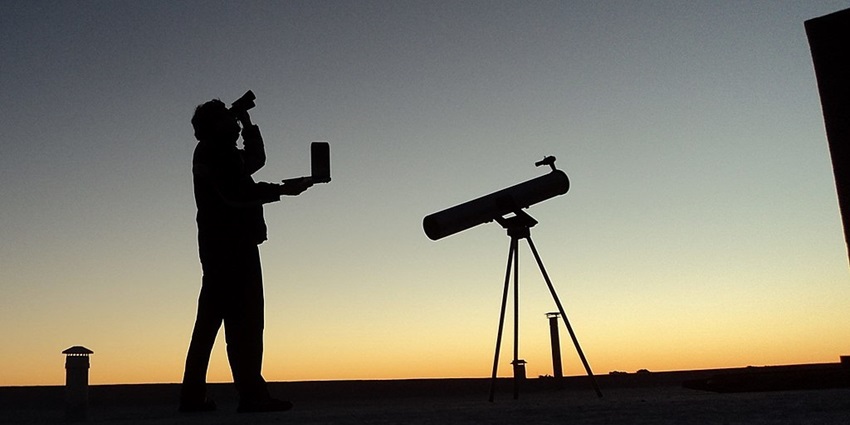 Silhouette of a man stargazing with his binoculars and a telescope on a tripod.