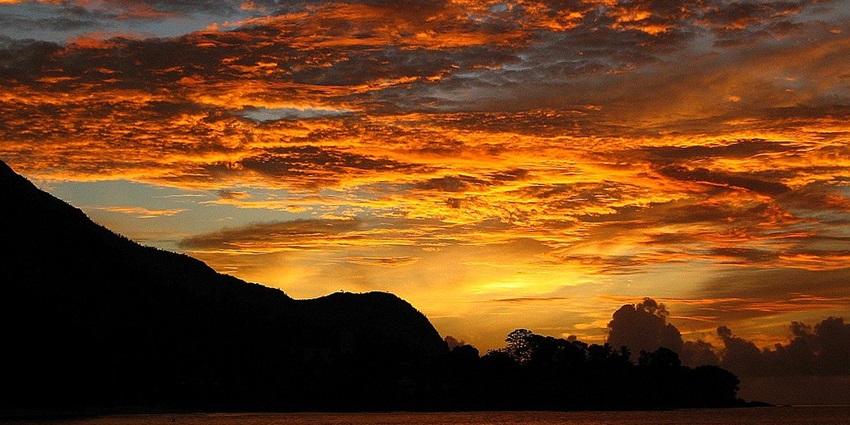 Sunset over a dark silhouette of a mountain and ocean, with orange and yellow clouds