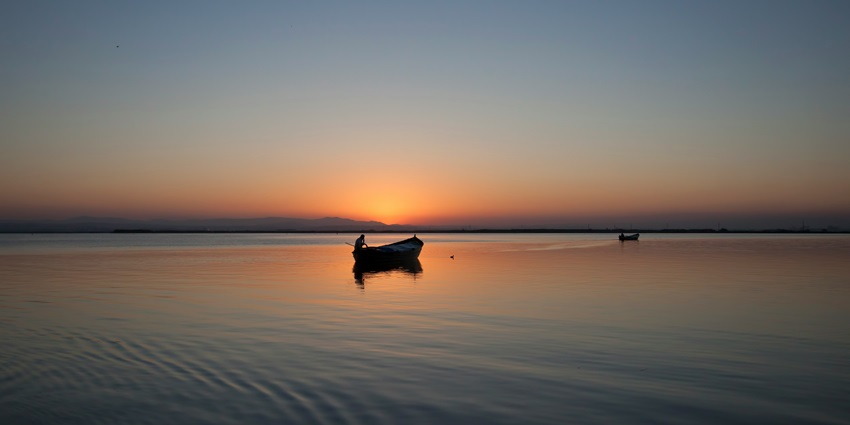 Person boating in the middle of a lake with the orange sky during sunset and distant buildings.