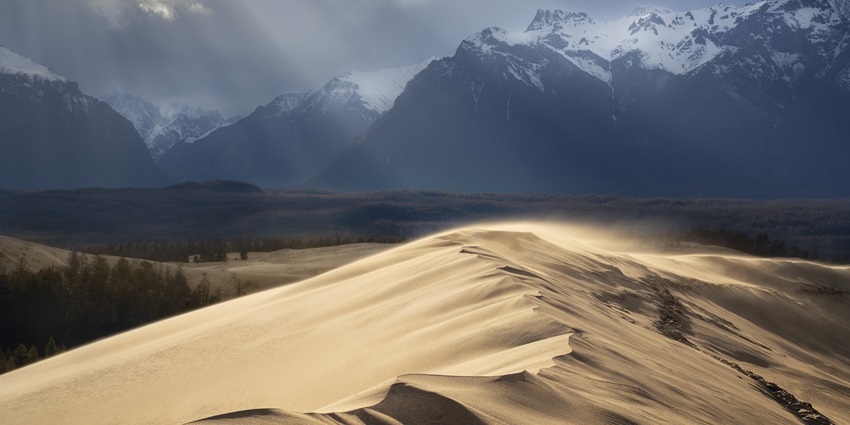 A large sand dune ridge with windblown sand, under a sky with sun rays breaking through