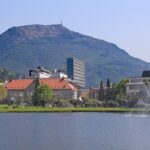 An image of Bergen, Norway, featuring a serene lake, fountains, buildings, and a mountain in the background.