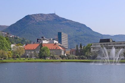 An image of Bergen, Norway, featuring a serene lake, fountains, buildings, and a mountain in the background.