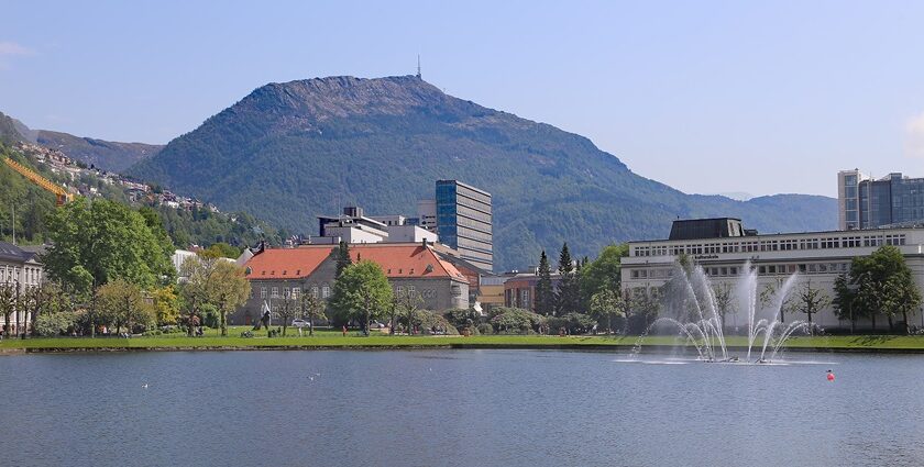 An image of Bergen, Norway, featuring a serene lake, fountains, buildings, and a mountain in the background.