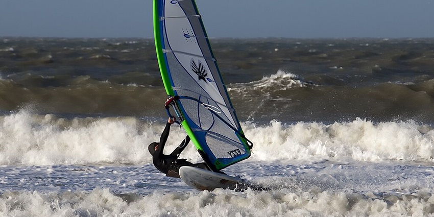 A windsurfer in a black wetsuit maneuvers a sailboard on a choppy ocean with large waves.