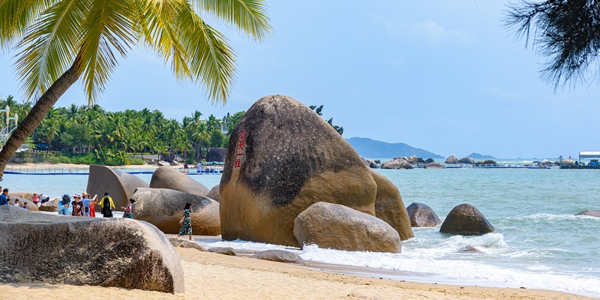 An image of Tianya Haijiao beach in China with a large boulder, clear water, and palm tree