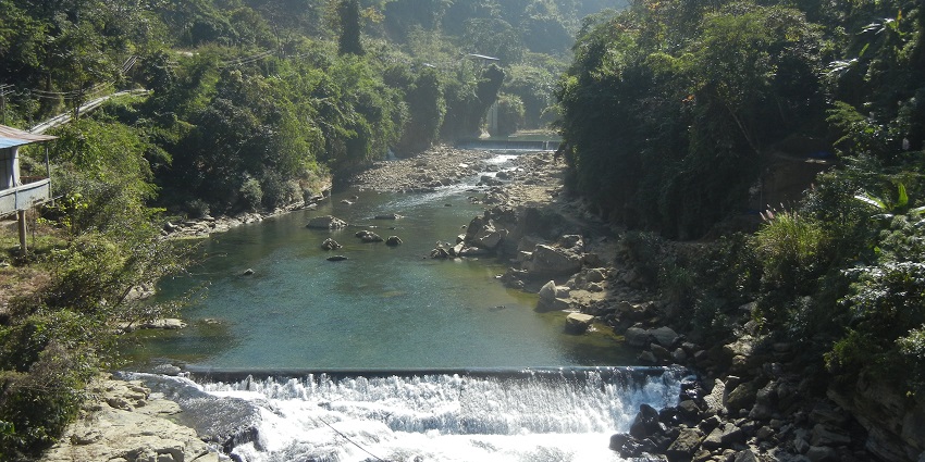 Tlawng River with rocks on both sides surrounded by dense greenery and a building