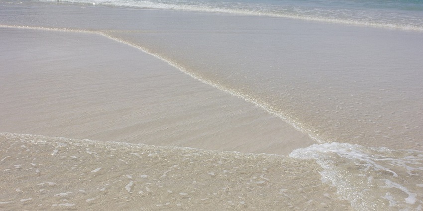 Shallow ocean waves gently lapping a pristine, light-colored sandy beach on a sunny day.