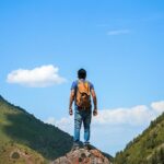 An image of a hiker wearing a backpack standing atop a rock, overlooking the greenery on mountains.