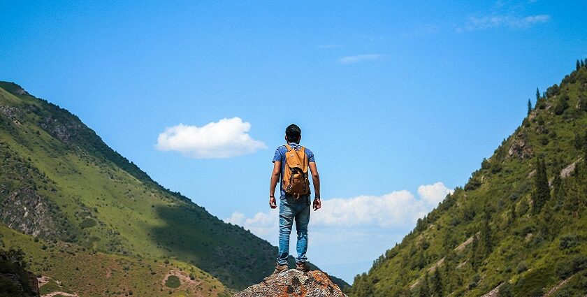 An image of a hiker wearing a backpack standing atop a rock, overlooking the greenery on mountains.