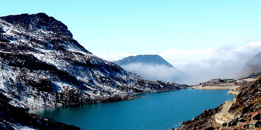 Tsomgo Lake with deep blue water, multiple buildings and hills partially covered in snow.