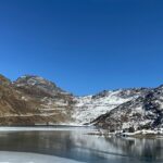 Tsomgo Lake with clear water surrounded by sandy, snowy shorelines and partial greenery