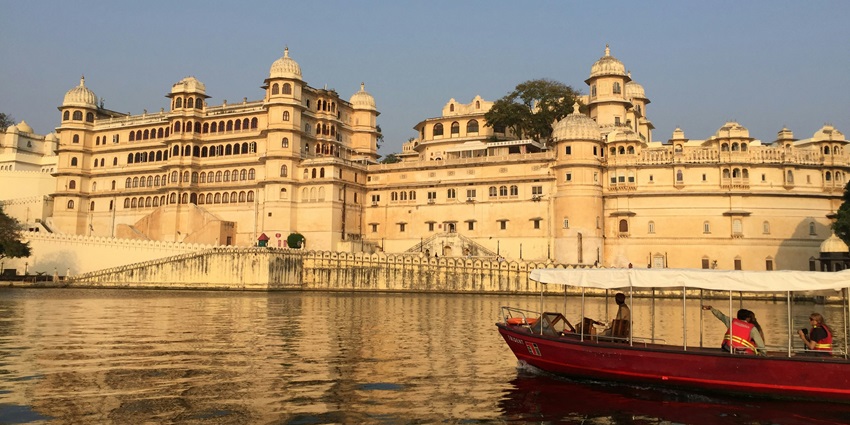 A large palace complex in Udaipur, with multiple buildings and trees, reflected in a calm lake.