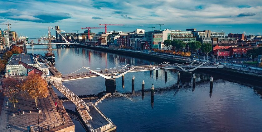 An Image of Dublin city featuring modern buildings, a river, and a bridge under a blue sky.