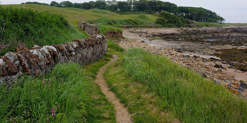 An image of the Coastal path with a stone wall overlooking a rocky beach and green hills.