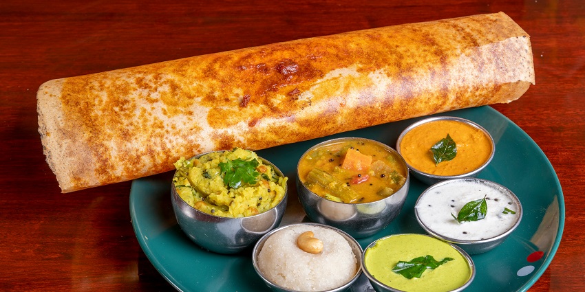 A crispy dosa served on a plate with a variety of chutneys, sambar, and potato filling.
