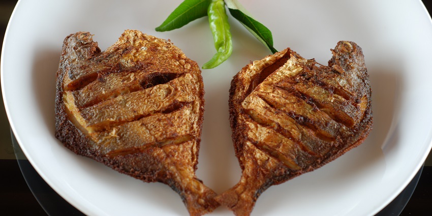 Karimeen fish fry served with rose-petaled tomato, green chilli and leaf on a white plate.