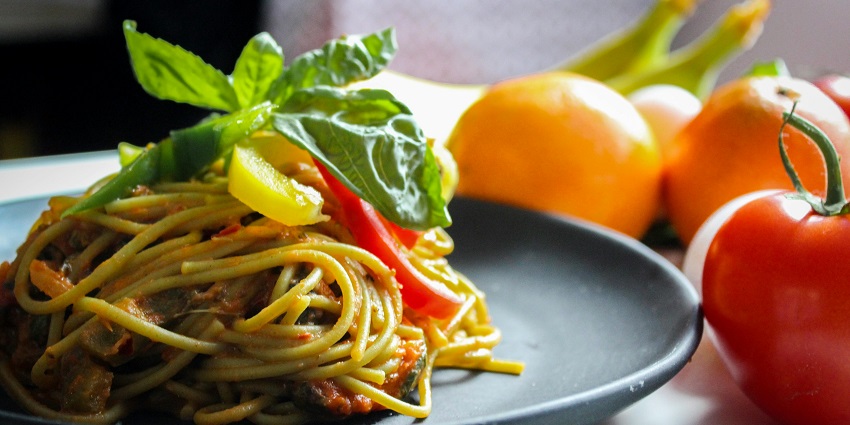 Image of a plate of Pasta With Vegetable Dish on Gray Plate Beside Tomato Fruit on White Table