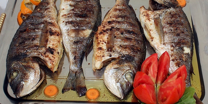 Four grilled fish, seasoned and cooked, arranged on a silver platter with orange slices